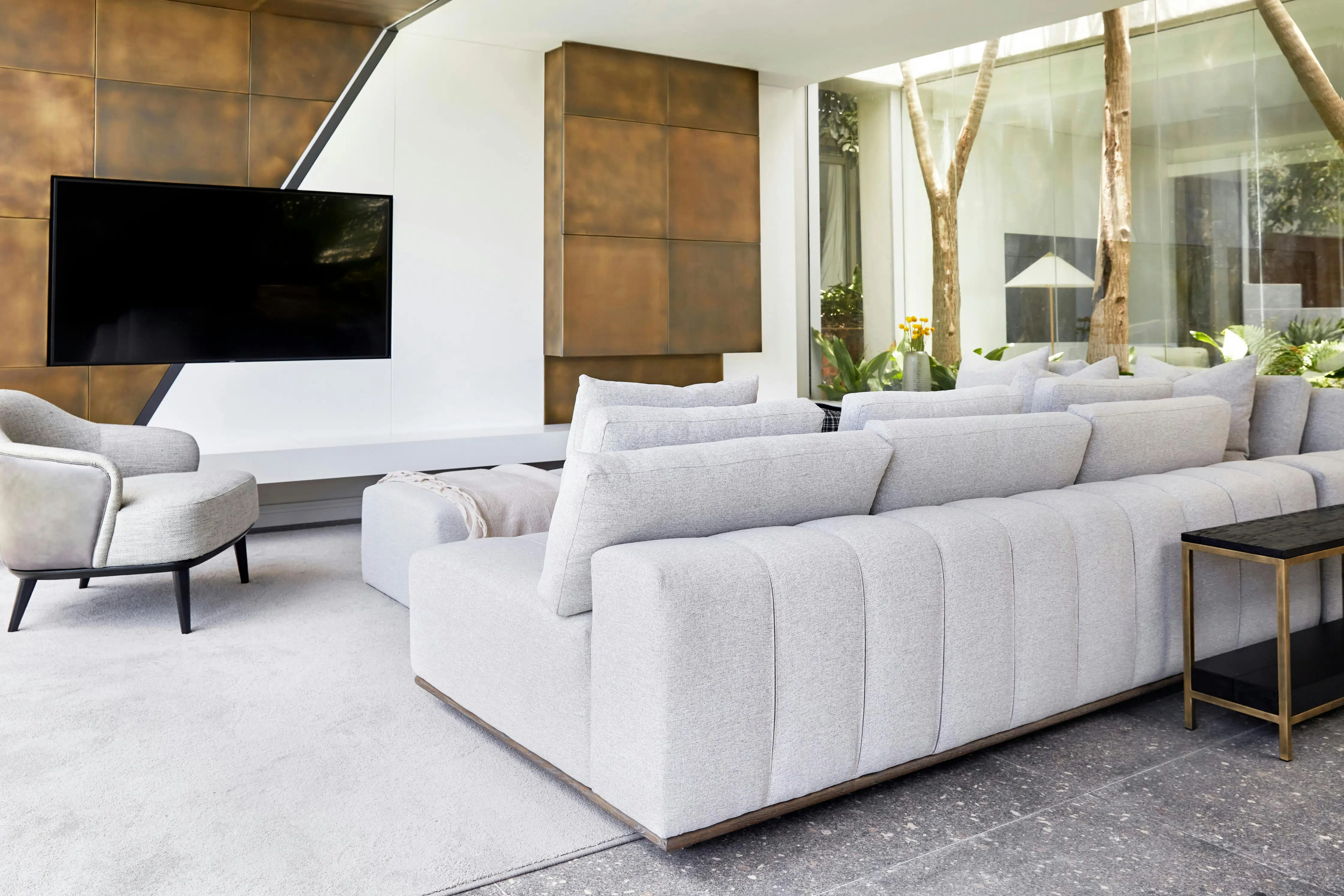 A minimalist living room with the back of a sofa in the foreground, a grey armchair diagonally to the left of the sofa, with a wall clad in antique brass plates in the background, adorned with a TV.