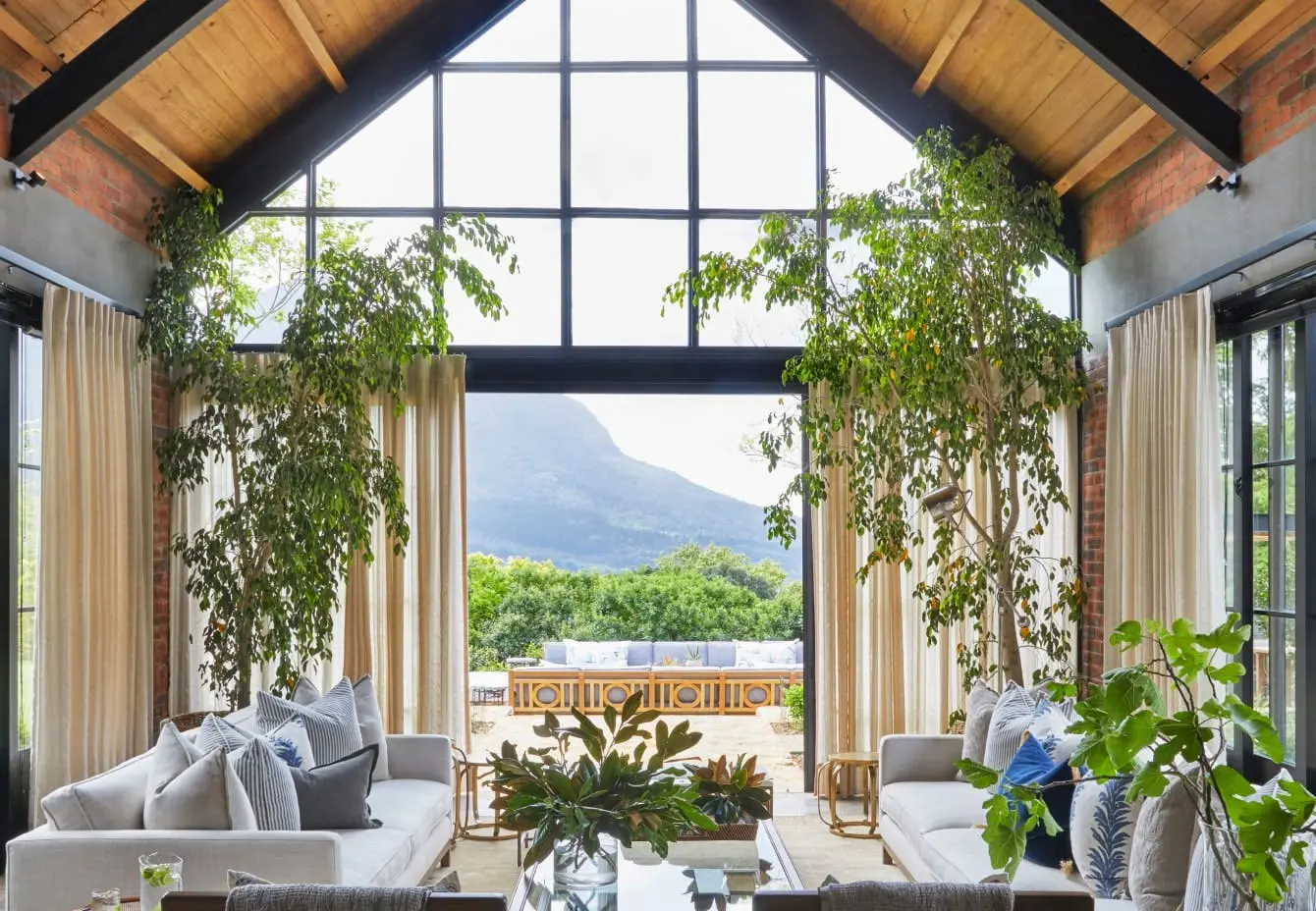 A living room with an a-frame roof featuring two occasional chairs and two sofas in the foreground with large plants that frame the door leading to the outside area in the background