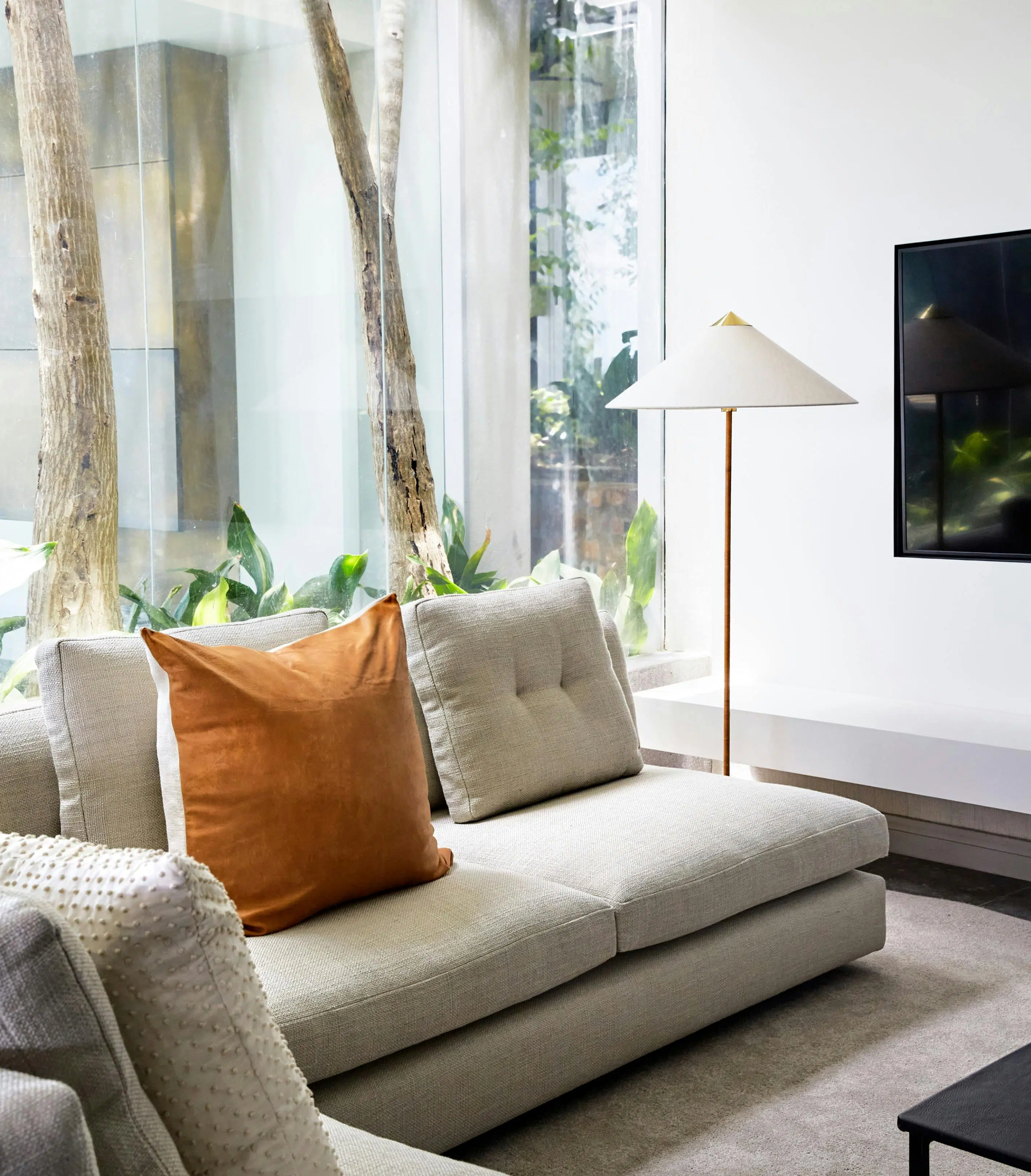A sofa with a modern floor lamp behind it with glass windows in the background looking onto two trees.