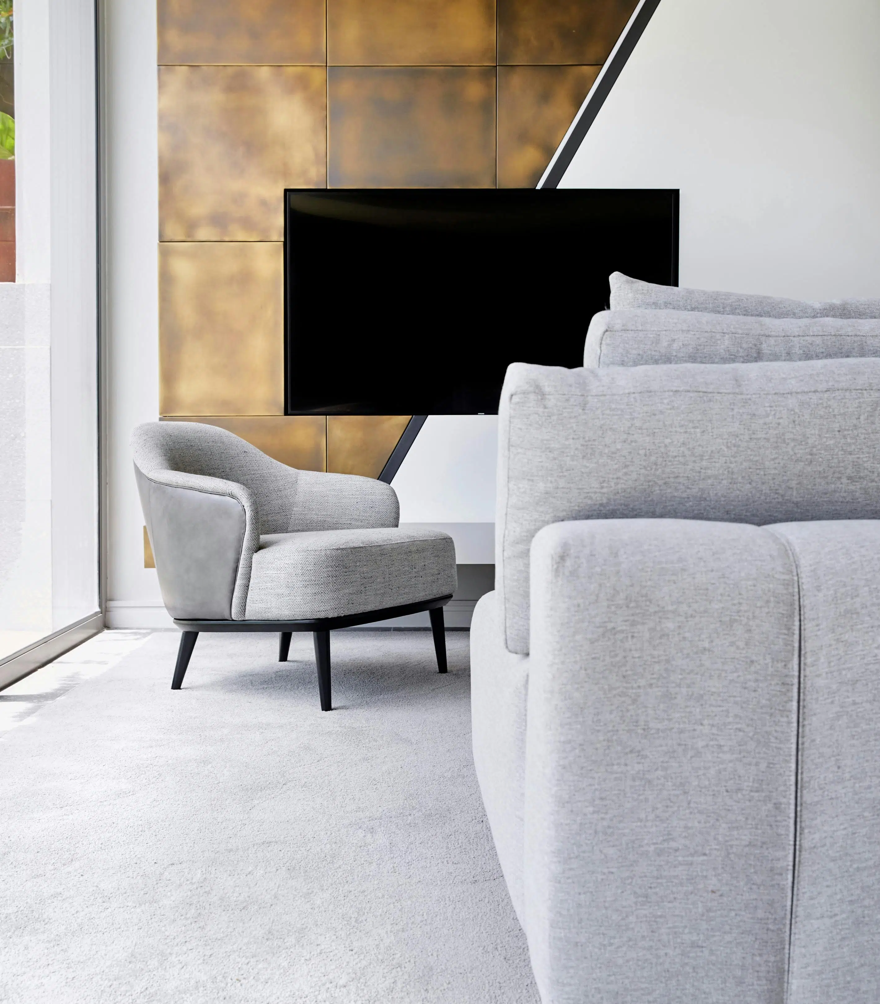 Close-up of a modern living room with a grey sofa in the centre of the image and a wall clad in antique brass plates in the background centred by a flat screen TV.
